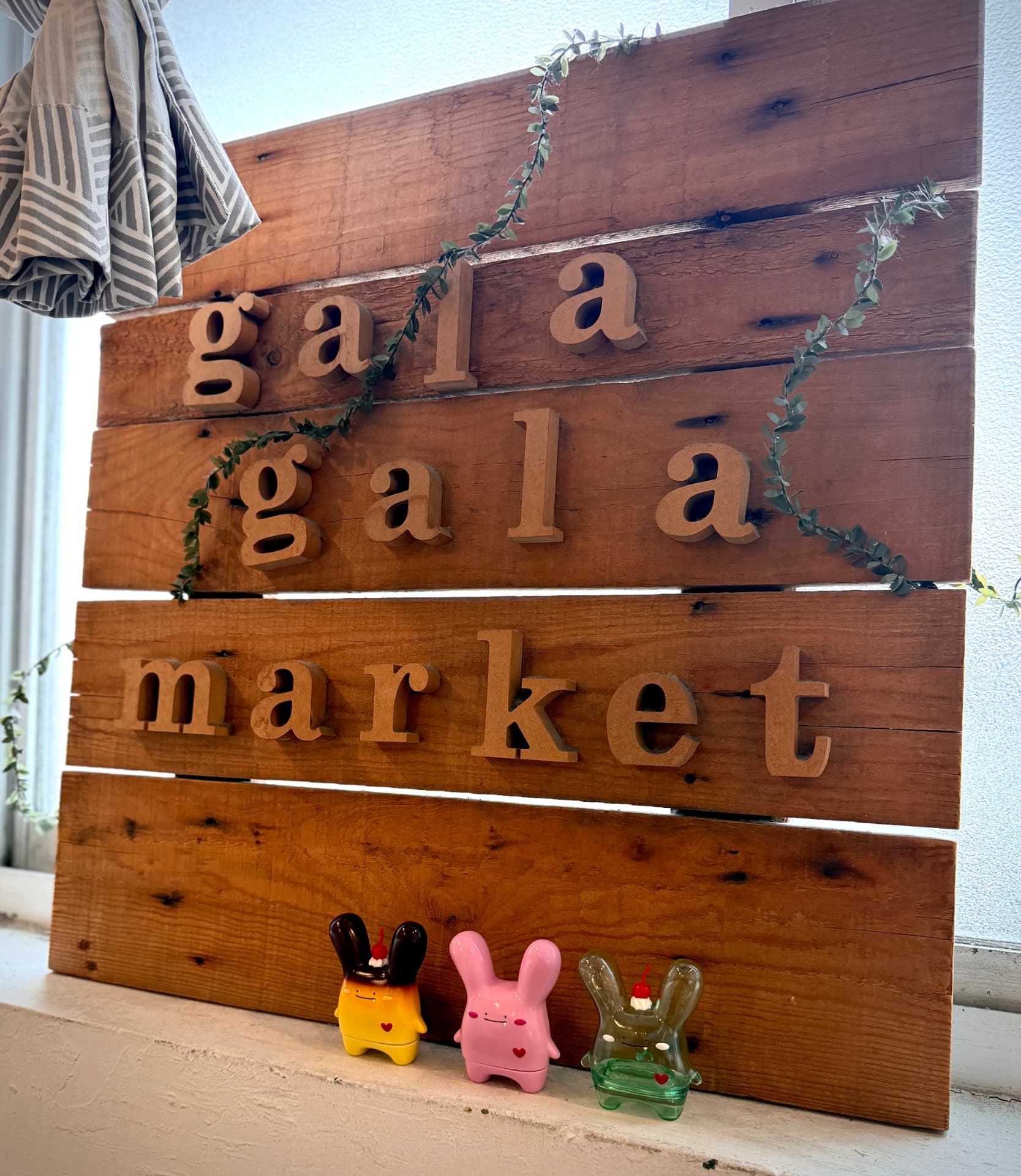 The wooden gala gala market plank sign on a windowsill, trailed with a eucalyptus vine and three Rabitt Planet figures sitting along the sill