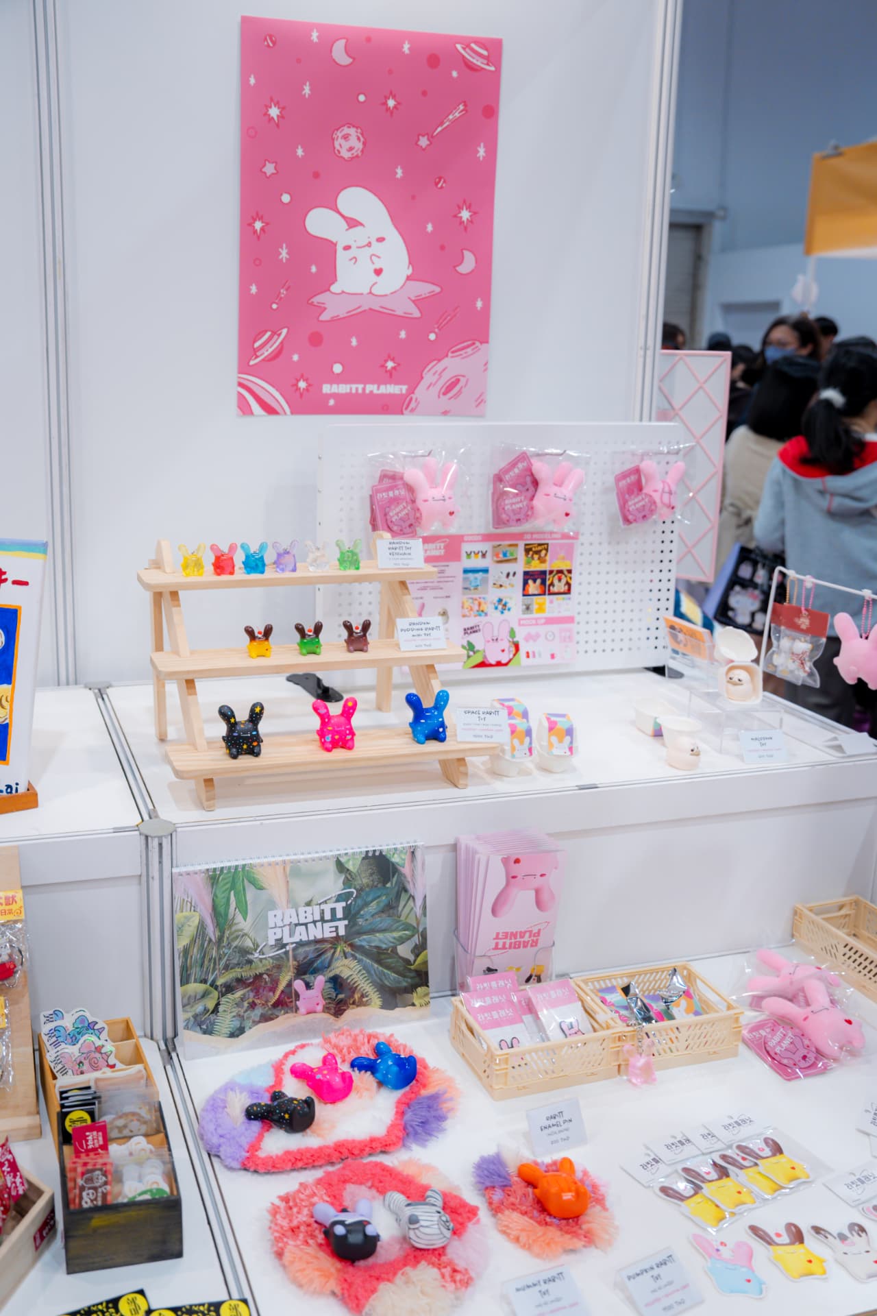 Rabitt Planet booth with a pink RABITT PLANET banner, packaged soft-vinyl rabbits and a wooden display of resin figures