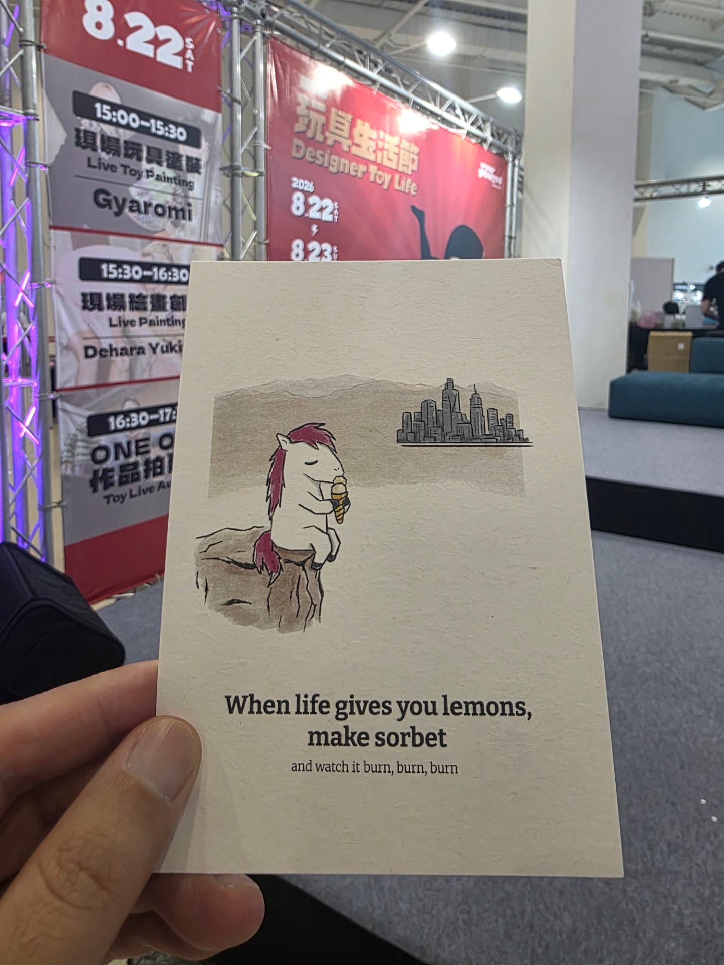 A hand holding an illustrated horse postcard at the Toy Life Festival in Kaohsiung, with event banners behind